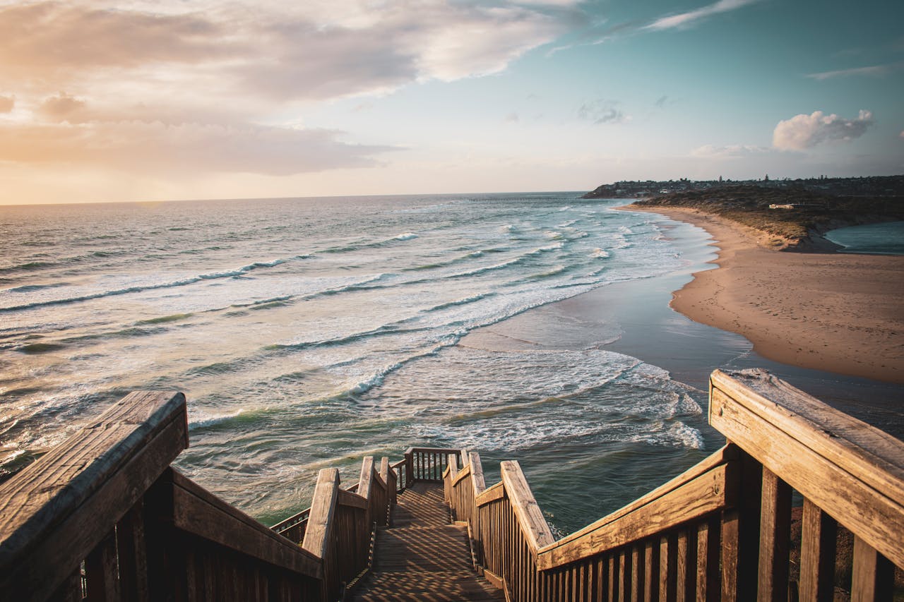 A picturesque view of the Adelaide beach with wooden stairs leading down, capturing waves and a sunset sky.